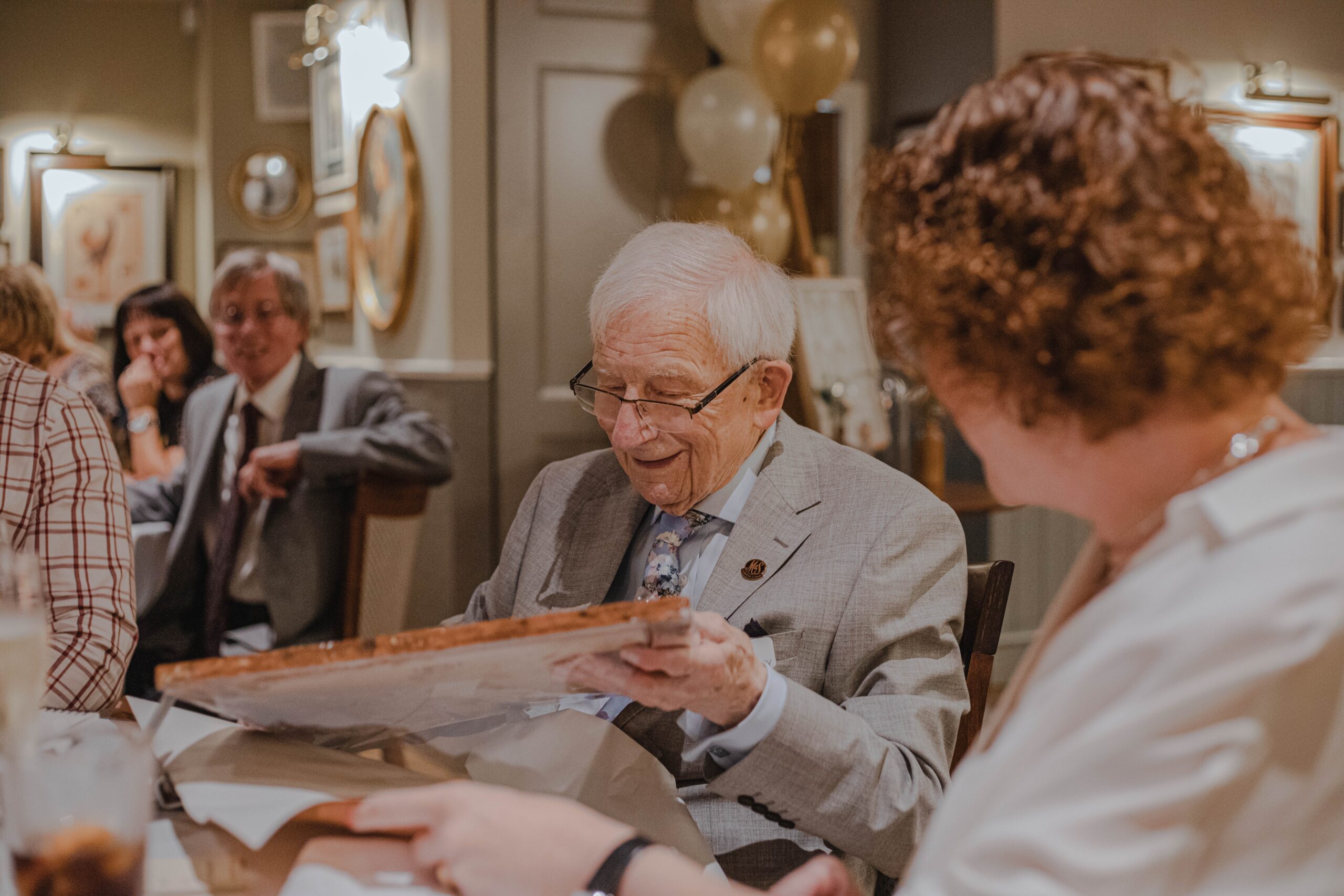 Choosing an aged care facility Smiling older man in a grey suit with a tie holding a picture in a room with other people